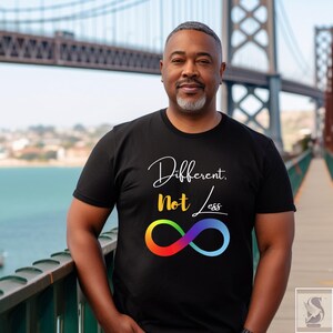 May include: Black t-shirt featuring the text "Different, Not Less" in white and yellow script, and a rainbow infinity symbol. The person is standing on a bridge.