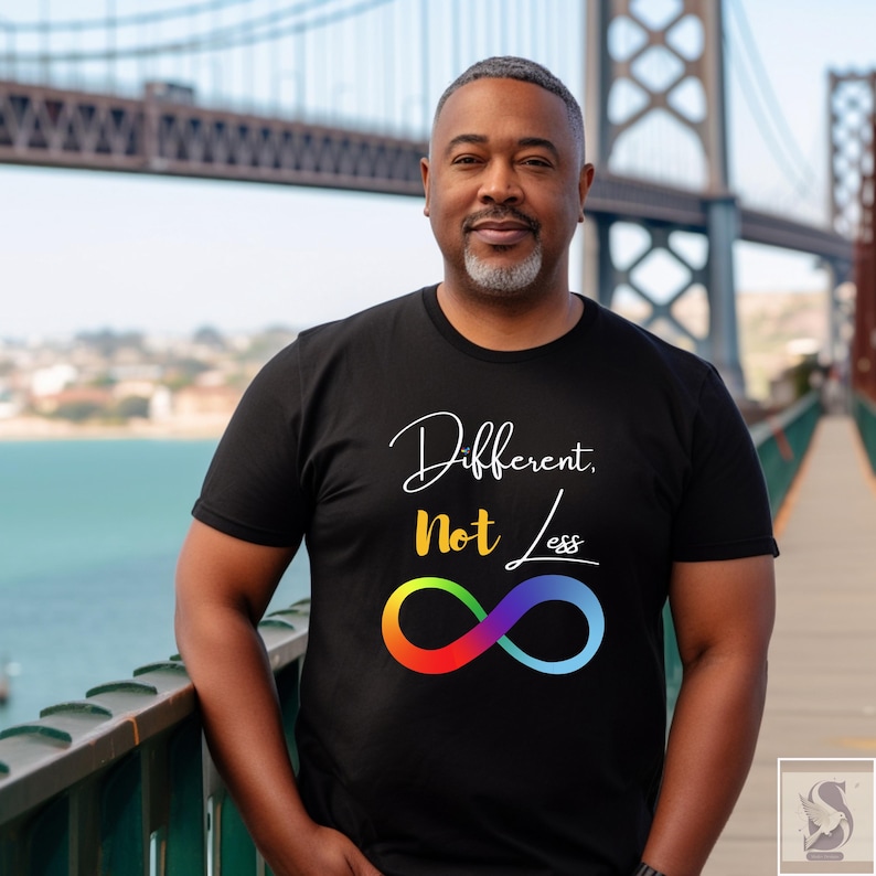 May include: Black t-shirt featuring the text "Different, Not Less" in white and yellow script, and a rainbow infinity symbol. The person is standing on a bridge.