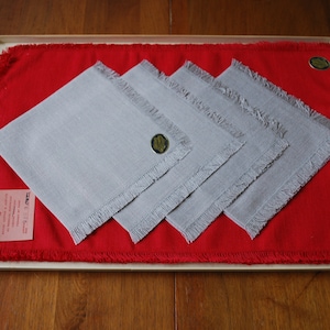 May include: Set of four gray linen napkins with fringed edges, displayed on a red linen placemat. The napkins are folded in half and have a small gold label with a design on each.