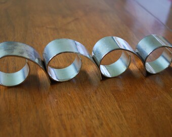 May include: Four silver napkin rings, each with a curved design, are arranged in a row on a wooden surface.