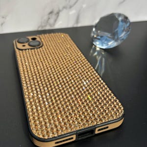 May include: A gold-colored phone case encrusted with small, sparkling rhinestones. The case has a black border and a tan-colored interior. A clear, faceted crystal is in the background.