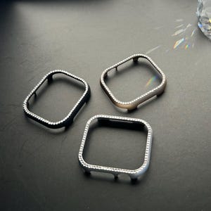 May include: Three watch case covers in black, silver, and gold, each encrusted with small, clear crystals. The cases are square with rounded corners and are displayed on a dark surface.