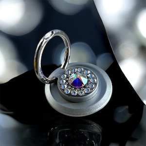 May include: A silver phone ring holder with a circular base and a ring for the finger. The base is adorned with a large, iridescent crystal surrounded by smaller clear crystals. The ring is attached to the base.