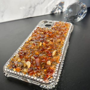 May include: A clear phone case bedazzled with small, amber-colored stones and clear rhinestones. The stones cover the back of the case, while the rhinestones line the edges. A large, clear, faceted crystal sits in the background.