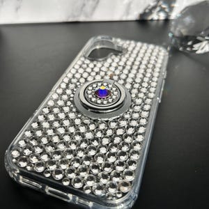 May include: Clear phone case covered in small, sparkling rhinestones. The case has a silver ring holder with a purple and blue center stone. The phone case is on a black surface.