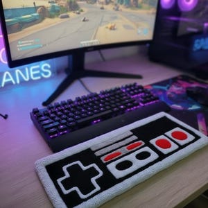 Könnte beinhalten: Ein schwarz-weißer Teppich in Form eines klassischen Videospiel-Controllers mit roten und grauen Akzenten. Eine schwarze Tastatur mit violetter Hintergrundbeleuchtung befindet sich hinter dem Teppich. Ein Computermonitor zeigt ein Videospiel.