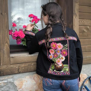 May include: A black denim jacket featuring a large, colorful embroidered floral bouquet on the back panel. The embroidery depicts pink, purple, and white flowers in a vase, framed by a decorative border. The cuffs also feature matching embroidery.