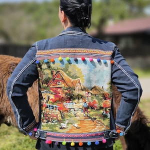 Pode incluir: Casaco de ganga azul escuro com um painel traseiro de tapeçaria que retrata uma cena rural. O casaco apresenta uma faixa decorativa com pompons multicoloridos na parte inferior e nos punhos. Costuras brancas.
