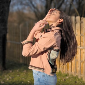 May include: A light pink jacket with brown trim and a decorative patch on the back, paired with light blue jeans. The jacket has long sleeves and a button-up front. The model is standing outside in front of a wooden fence.