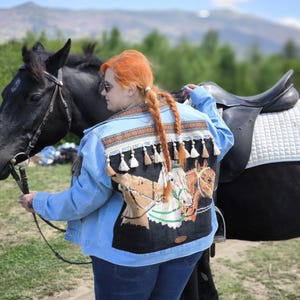 May include: Light blue denim jacket with a detailed horse-themed design on the back. The design features horses in shades of brown and white, with decorative tassels and trim. The jacket is paired with blue jeans.