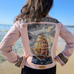 May include: A light pink jacket featuring a large rectangular needlepoint panel on the back depicting a sailing ship on the ocean under a cloudy sky. The jacket includes decorative patterned trim on the shoulders and cuffs, with a small tan label.