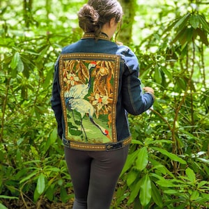 Pode incluir: Jaqueta jeans azul com um painel traseiro de tapeçaria detalhado com um desenho de garça e floral em tons de verde, marrom e branco. A jaqueta é de cor azul clássica e tem fecho de botões.