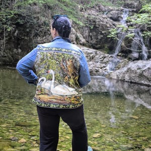 Puede incluir: Chaqueta vaquera azul claro con una pintura detallada de dos cisnes blancos en un paisaje lacustre. La chaqueta tiene un borde decorativo. La persona está cerca de una cascada.