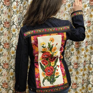 以下が含まれることがあります： 背中にカラフルな花の刺繍が施された濃いブルーのデニムジャケット。刺繍は赤いポピー、黄色とオレンジの花、緑の葉が特徴です。ジャケットは肩と袖口に装飾的なトリムが付いています。