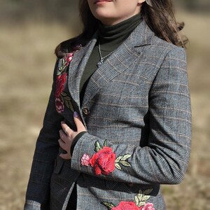 May include: A gray plaid blazer with red floral embroidery on the lapel and sleeve. The blazer is buttoned and worn over a dark green turtleneck. The person is wearing blue jeans.