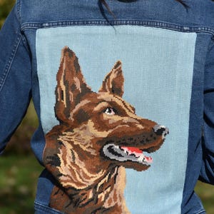 May include: A blue denim jacket with a large embroidered patch on the back. The patch features a detailed portrait of a German Shepherd dog in shades of brown and tan against a light blue background.