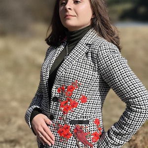 May include: A black and white houndstooth blazer featuring a red floral and bird embroidery. The jacket has a classic design with a notched lapel and two front pockets. The model is wearing a green turtleneck and blue jeans.