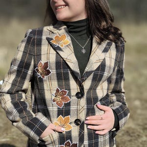 Op de afbeelding: Geruite blazer in beige, zwart en bruin met bladeren applicaties. De jas heeft een klassiek ontwerp met een reverskraag, twee knopen en zakken. De bladeren zijn in bruin- en goudtinten. De persoon draagt een donkergroene coltrui.