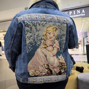 May include: A blue denim jacket with a colorful embroidered border and a large embroidered patch on the back depicting a girl with cats. The patch is a tapestry-style design with a light blue background and a girl in a pink dress holding a cat.