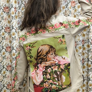 Puede incluir: Chaqueta beige con bordados florales en hombros y mangas. Un gran parche bordado en la espalda presenta a una mujer de pelo largo, apoyada en una mesa con flores. La chaqueta es de color beige claro.