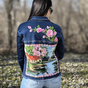 May include: A blue denim jacket with a floral embroidery design on the back. The embroidery features a pink and green landscape with a house, trees, and a river.