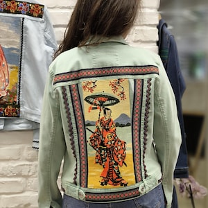Può includere: Giacca di jeans verde chiaro con un pannello ricamato dettagliato sul retro raffigurante una geisha giapponese con un ombrello. Il ricamo include dettagli arancioni, rossi e neri. La giacca ha una chiusura con bottoni ed è abbinata a jeans blu.