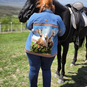 May include: A denim jacket with a horse-themed design on the back, paired with blue jeans. The jacket features a colorful image of running horses. A black horse with a saddle stands nearby on a grassy field.