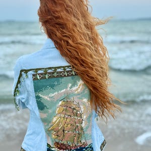 May include: A light blue denim jacket featuring a large rectangular needlepoint panel on the back depicting a sailing ship on the ocean. The jacket is accented with a decorative green and brown patterned trim along the hem and shoulders.