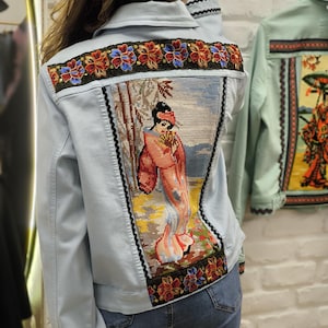 Peut inclure: Veste en jean bleu clair avec broderie complexe au dos représentant une femme en costume traditionnel. La veste présente des motifs floraux et géométriques rouges, bleus et dorés. Bordure noire.