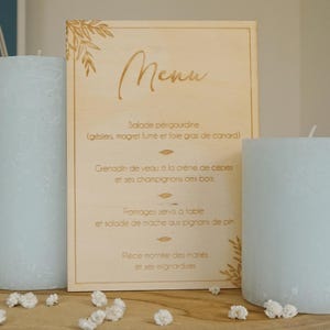 Puede incluir: Un letrero de menú de madera con una inscripción dorada. El menú incluye una ensalada, un plato de ternera, quesos y un pastel de boda. El letrero está sobre una mesa con flores blancas.
