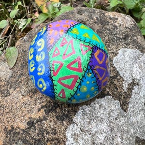 May include: A hand-painted, colourful rock with a patchwork design. The rock features sections of blue, green, purple, and orange, each with unique patterns and shapes. Red triangles and yellow dots add detail. The rock is sitting on a larger rock.