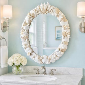 May include: An oval wall mirror featuring a frame densely decorated with various white and cream-colored seashells, coral pieces, and sand dollars. The mirror is mounted on a light blue wall above a white marble vanity countertop with a sink.