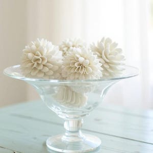 May include: A clear glass pedestal bowl filled with several white, spherical decorative flowers. The bowl sits on a light blue, distressed wooden table. The background is a soft, blurred view of a window and a white wall.