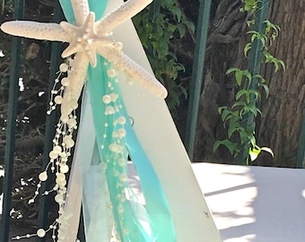 May include: White wooden chair decorated with a large white starfish, turquoise ribbons, and strings of white pearls. The chair is set up for a beach-themed event, with a natural background.