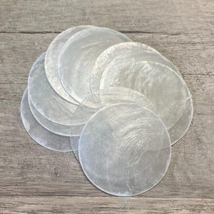 May include: A collection of iridescent, round, mother-of-pearl shell discs. The discs are a pearlescent white and arranged in a slightly overlapping pattern. The natural material has a subtle, textured surface, reflecting light.