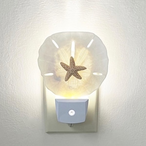 May include: A white night light shaped like a sand dollar with a starfish inside. The light is plugged into an electrical outlet.