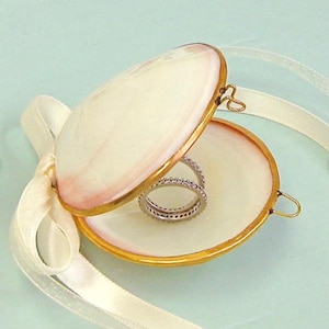 Puede incluir: Una caja de anillo de concha de almeja blanca con un borde dorado y una cinta de satén blanca. La caja está abierta y hay dos anillos de plata con pequeños diamantes dentro.
