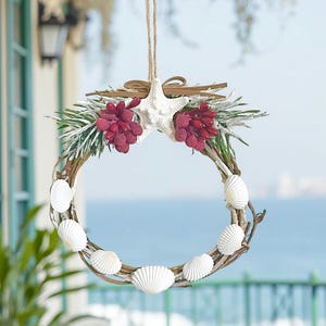 May include: A decorative wreath featuring a circular grapevine base adorned with white seashells, red berries, and a white starfish. The wreath is accented with green sprigs and a brown bow, suspended by a natural twine.