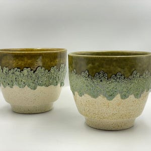 May include: Two ceramic planters with a layered design. The top is a dark olive green, transitioning to a light green, and then a speckled beige at the base. The planters have a textured, dripped glaze effect.
