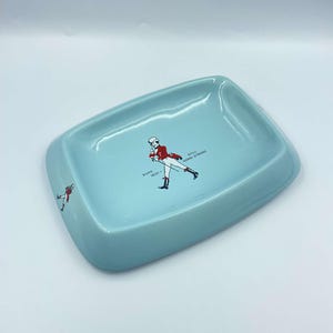 May include: A light blue ceramic ashtray featuring a graphic of a man in a red jacket and black trousers walking with a stick. The text "BORN 1820 - STILL GOING STRONG" is printed on the ashtray.