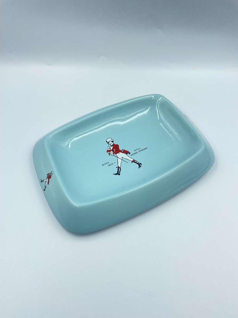 May include: A light blue ceramic ashtray featuring a graphic of a man in a red jacket and black trousers walking with a stick. The text "BORN 1820 - STILL GOING STRONG" is printed on the ashtray.