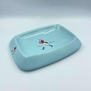 May include: A light blue ceramic ashtray with a white outline and a graphic of a man in a red jacket and white trousers walking with a cane. The text "BORN 1820" and "STILL GOING STRONG" is printed on the ashtray.