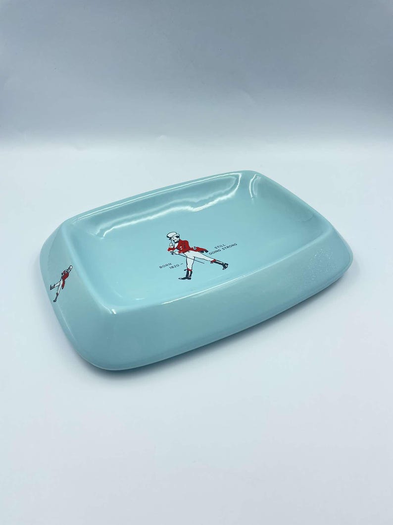 May include: A light blue ceramic ashtray with a white outline and a graphic of a man in a red jacket and white trousers walking with a cane. The text "BORN 1820" and "STILL GOING STRONG" is printed on the ashtray.