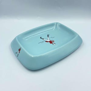 May include: A light blue ceramic ashtray with a white design featuring a man in a red coat and black trousers walking with a cane. The text "SMOKES DRUNK THIS - OVER -  YOUR -  MEASURE" is printed on the ashtray.