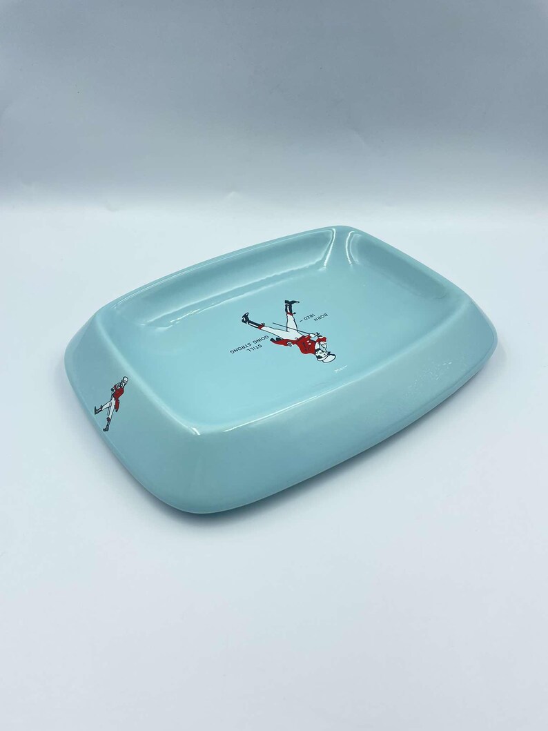 May include: A light blue ceramic ashtray with a white design featuring a man in a red coat and black trousers walking with a cane. The text "SMOKES DRUNK THIS - OVER -  YOUR -  MEASURE" is printed on the ashtray.