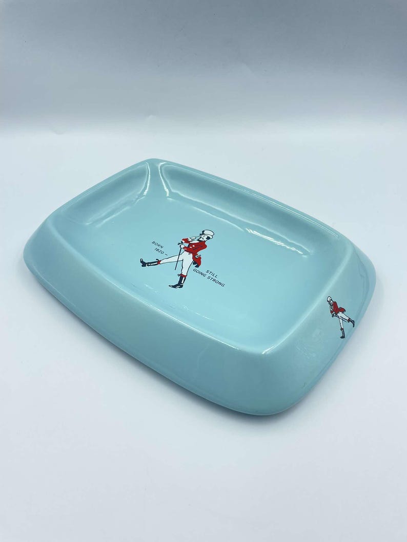 May include: A light blue ceramic ashtray featuring a walking man graphic and the text "Born 1820 - Still Going Strong".