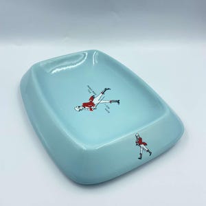 May include: A light blue ceramic ashtray with a white and red illustration of a man in a red jacket and white trousers. The text "BORN 1820" is printed on the ashtray.