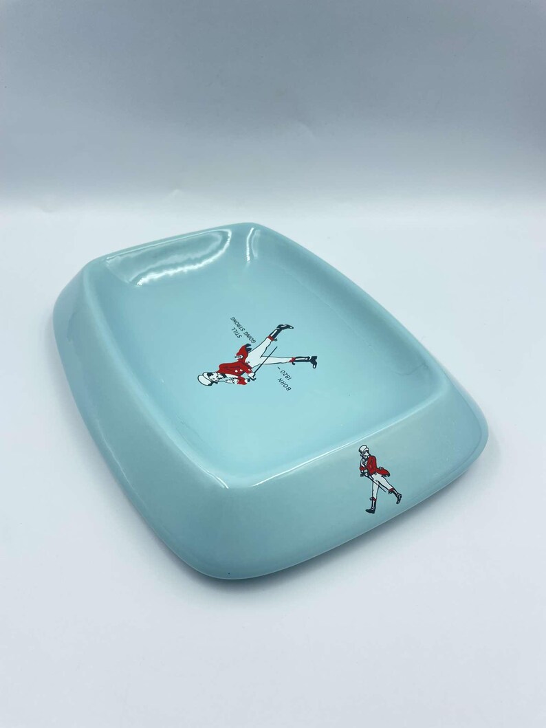 May include: A light blue ceramic ashtray with a white and red illustration of a man in a red jacket and white trousers. The text "BORN 1820" is printed on the ashtray.