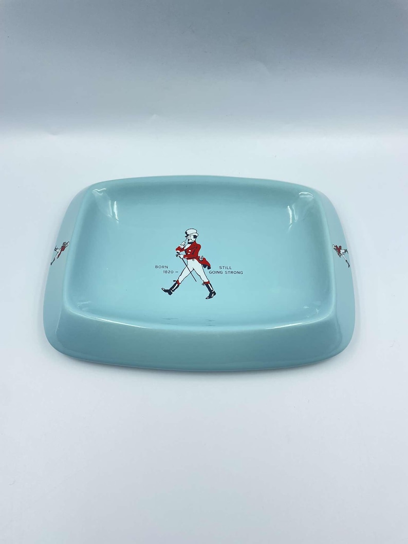 May include: A light blue ceramic ashtray featuring a walking man graphic and the text "Born 1820 - Still Going Strong".