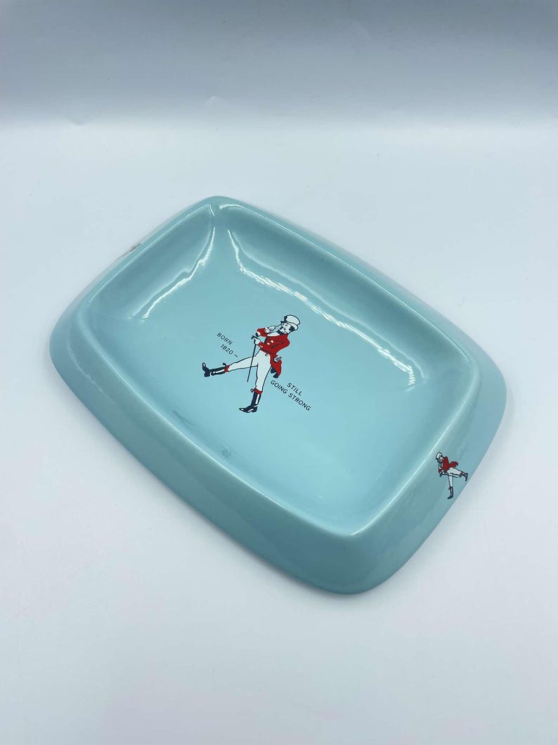 May include: A light blue ceramic ashtray featuring a red and white illustration of a man in riding attire. The text "BORN 1820 - STILL GOING STRONG" is printed on the ashtray.